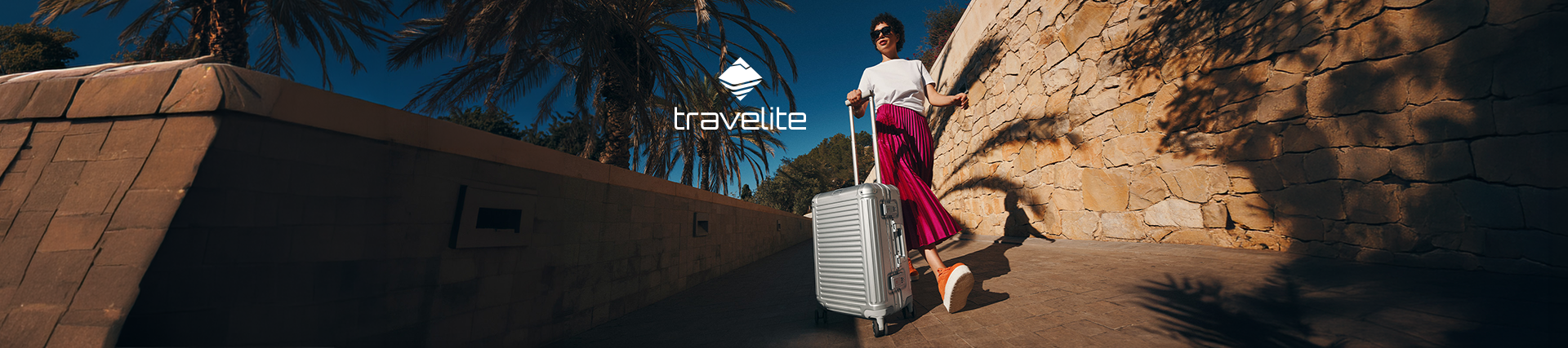 Frau zieht einen silbernen Travelite Hartschalenkoffer durch eine sonnige mediterrane Umgebung mit Palmen und Natursteinmauer.