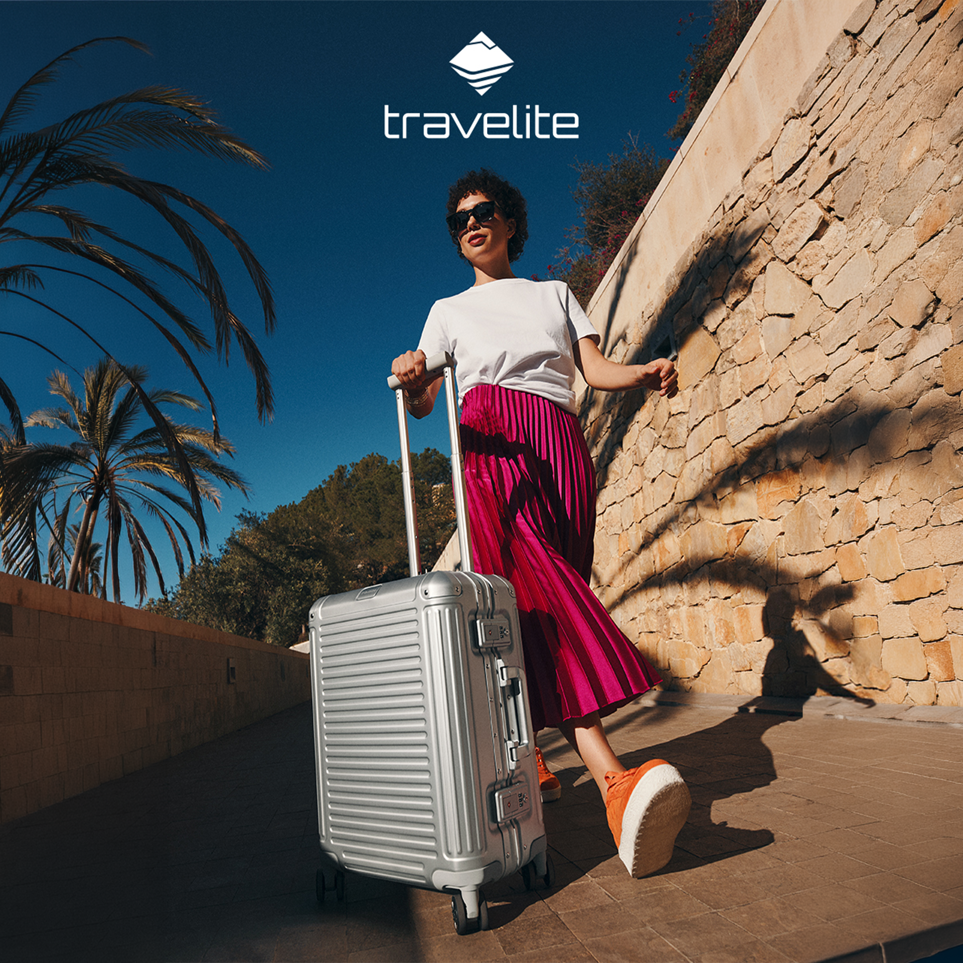 Frau mit Sonnenbrille zieht einen großen silbernen Travelite Hartschalenkoffer an Palmen und einer Natursteinmauer entlang.
