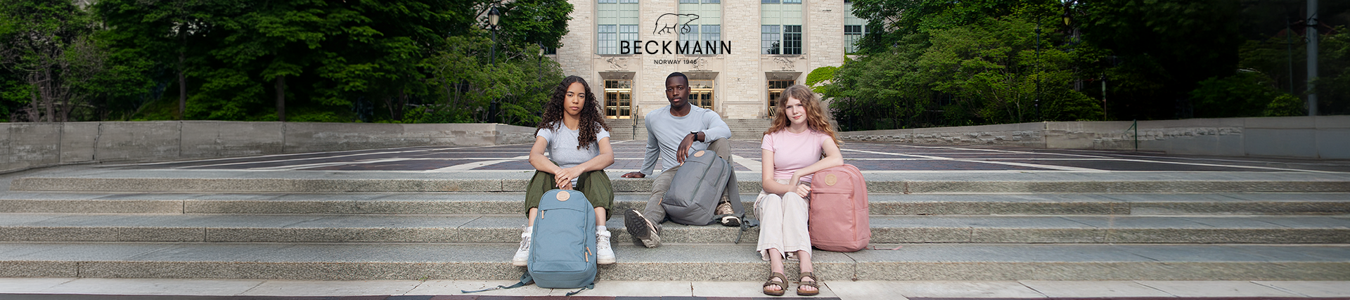 Drei junge Erwachsene sitzen mit Beckmann Rucksäcken in Blau, Grau und Rosa auf den Stufen eines Universitätsgebäudes.